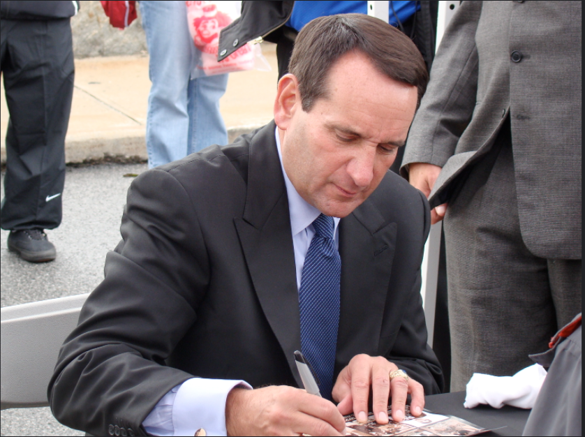 Sitting down at a table at the Army football stadium Coach K signs autographs for fans. He was a great coach and will be remembered for his great successes.